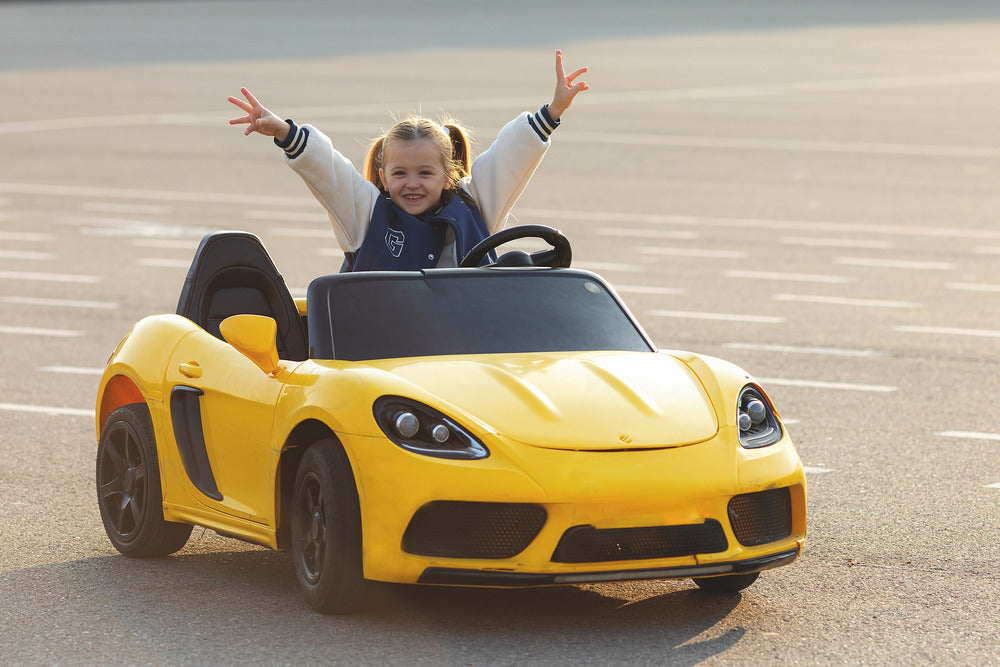 A little girl in a varsity jacket sits behind the wheel of her yellow, electric, ride-on sports car.