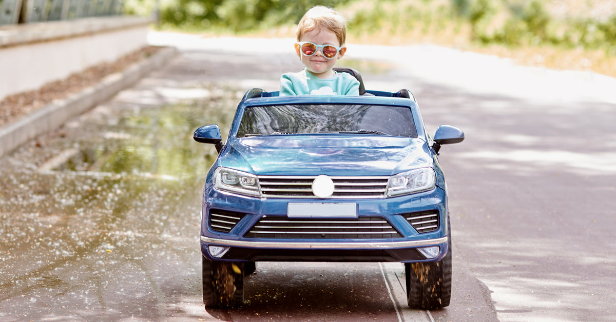 A little girl wears sunglasses while driving a blue power wheel over a bridge. A forest stands behind her.