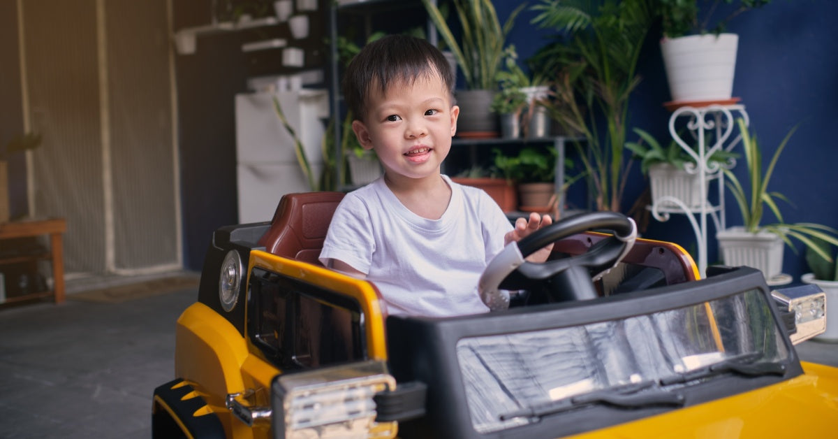 A young boy sits behind the wheel of his yellow ride-on car. He is sitting in his house with plants in the background.